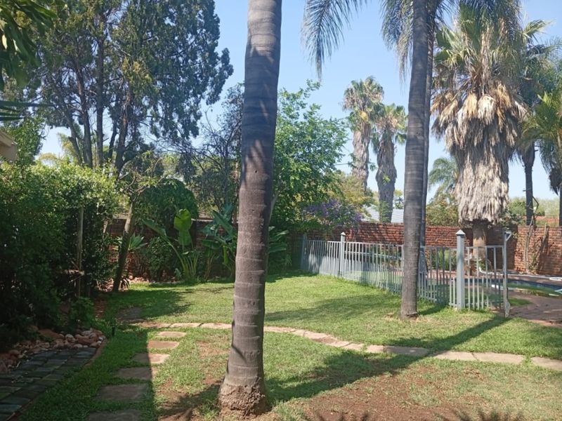 A private garden area featuring a lush lawn, mature palm trees, stone pathways, and a secure metal fence enclosing the pool area.