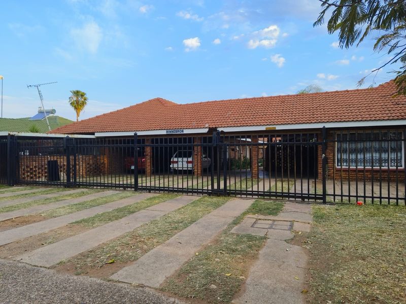 Front elevation of a brick apartment complex featuring a tiled roof, secure metal perimeter fencing, and a paved driveway with carport parking.