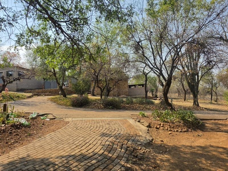 A spacious farm garden area featuring mature trees, a brick-paved walkway, and rustic stone masonry outbuildings set within a natural landscape.