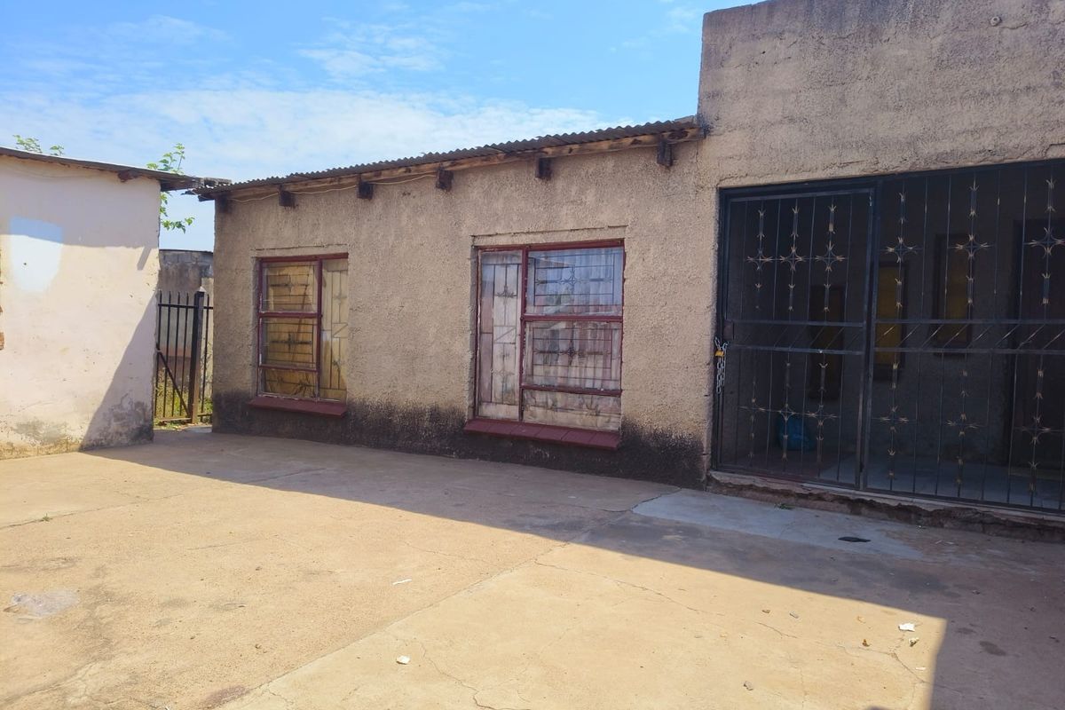 Rear exterior view of the property featuring a concrete paved yard, security gates, and weathered wall finishes.