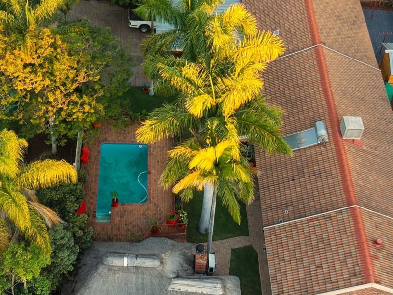 Aerial view of the property featuring a swimming pool, brick-paved patio area, mature landscaping, and a tiled roof with a solar water heating system.