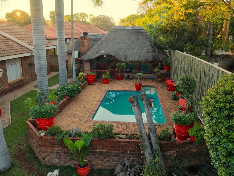 An elevated view of a backyard featuring a swimming pool, brick-paved patio, and a traditional thatched lapa entertainment area.