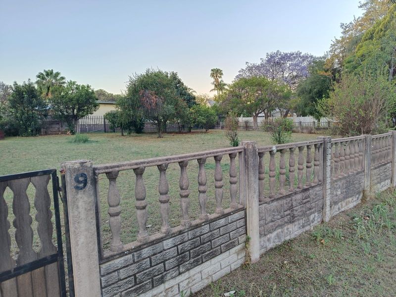 A vacant residential plot featuring a mature garden, established trees, and a precast concrete boundary fence with a metal pedestrian gate.