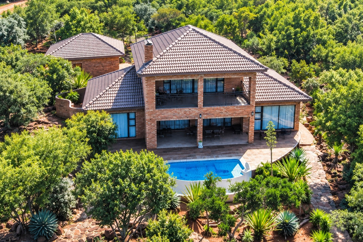 Aerial view of a two-story brick house featuring a tiled roof, balcony, covered patio, and a swimming pool set within a lush, wooded landscape.