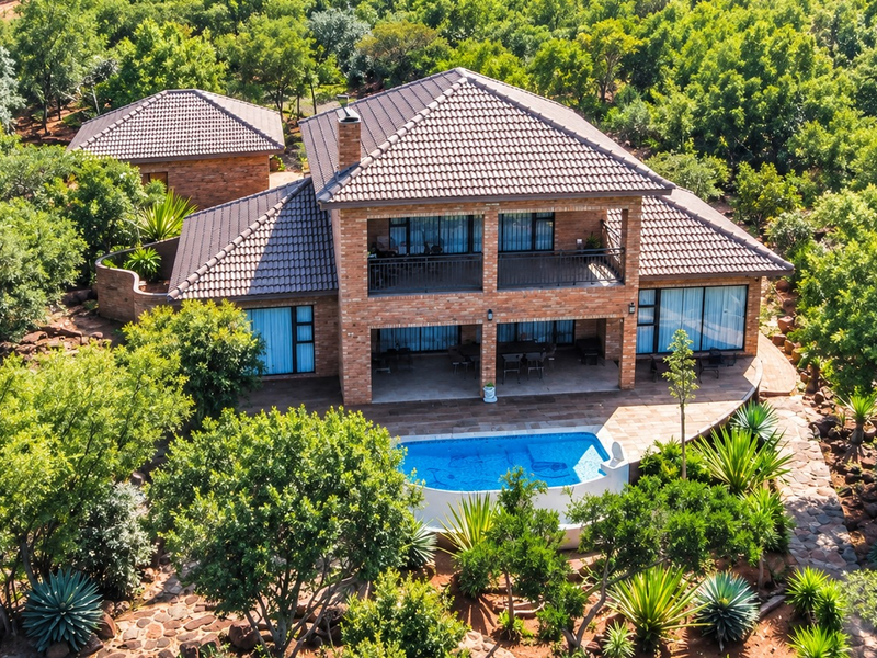 Aerial view of a two-story brick house featuring a tiled roof, balcony, covered patio, and a swimming pool set within a lush, wooded landscape.