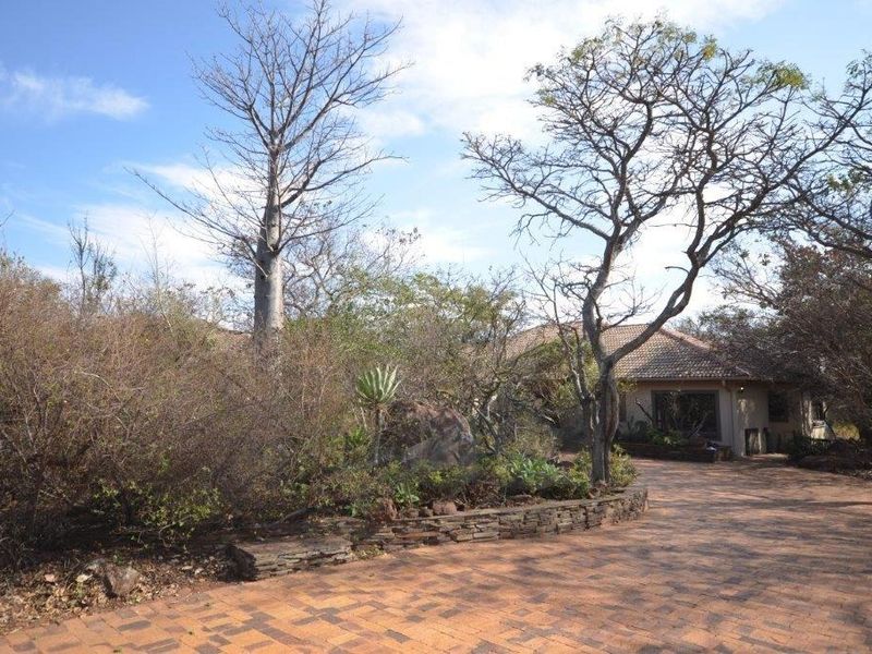 Front exterior view of the property featuring a brick paved driveway, stone retaining wall, and established garden with mature trees.