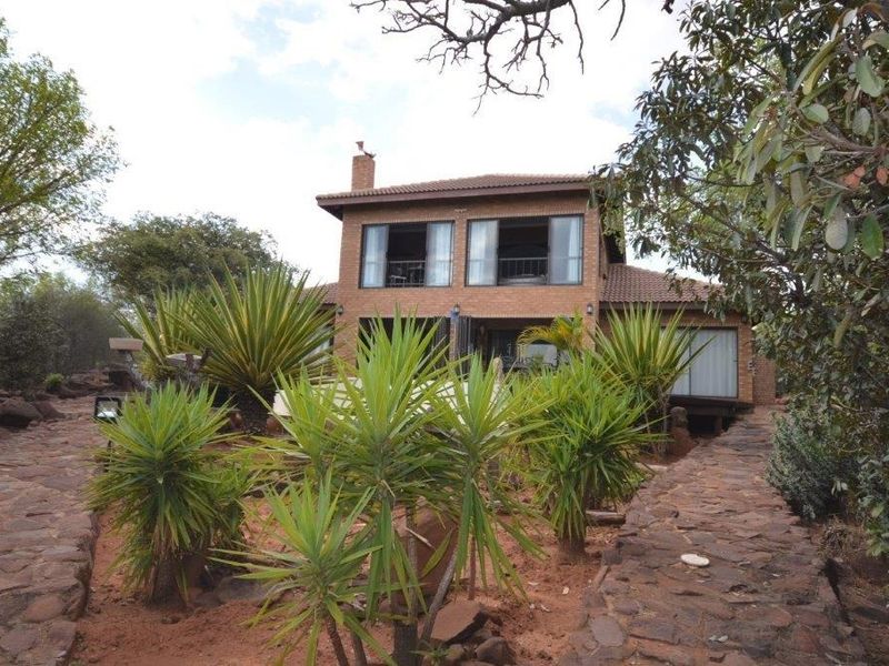 A two-story brick house featuring a tiled roof, chimney, and sliding glass doors, set within a landscaped garden with stone pathways.