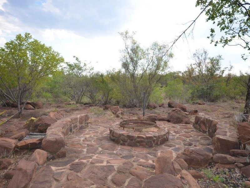 An outdoor garden area featuring a rustic stone-paved patio with a built-in fire pit and curved stone seating, set within a natural bushveld landscape.