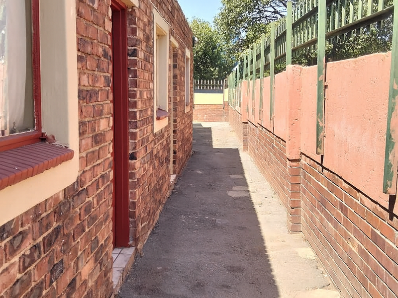 A narrow exterior side passage featuring face brick walls, a paved walkway, and a perimeter wall with metal security fencing.