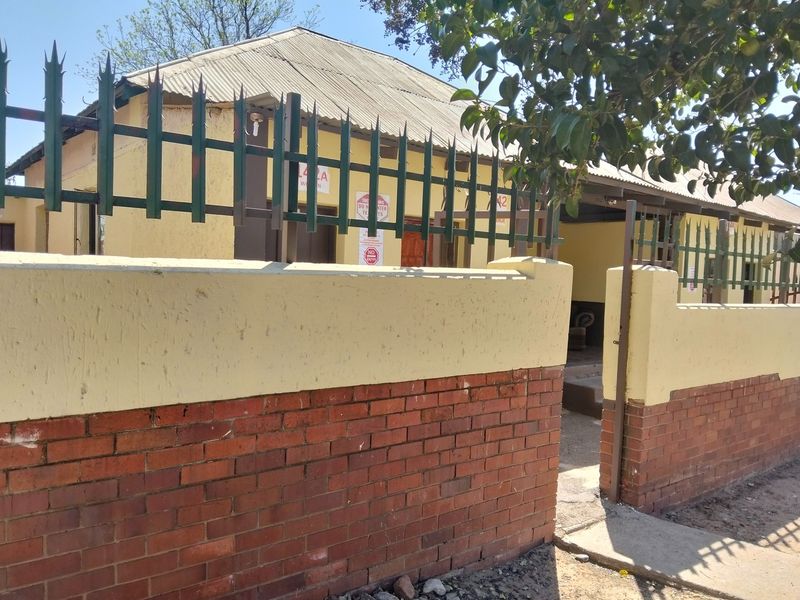 Front exterior view of a single-story house featuring a corrugated iron roof, brick boundary wall, and metal security fencing.