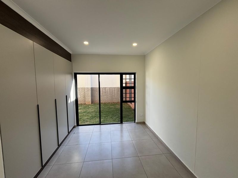 A bright, empty bedroom featuring modern built-in cupboards, large sliding glass doors leading to a garden, and neutral tiled flooring.