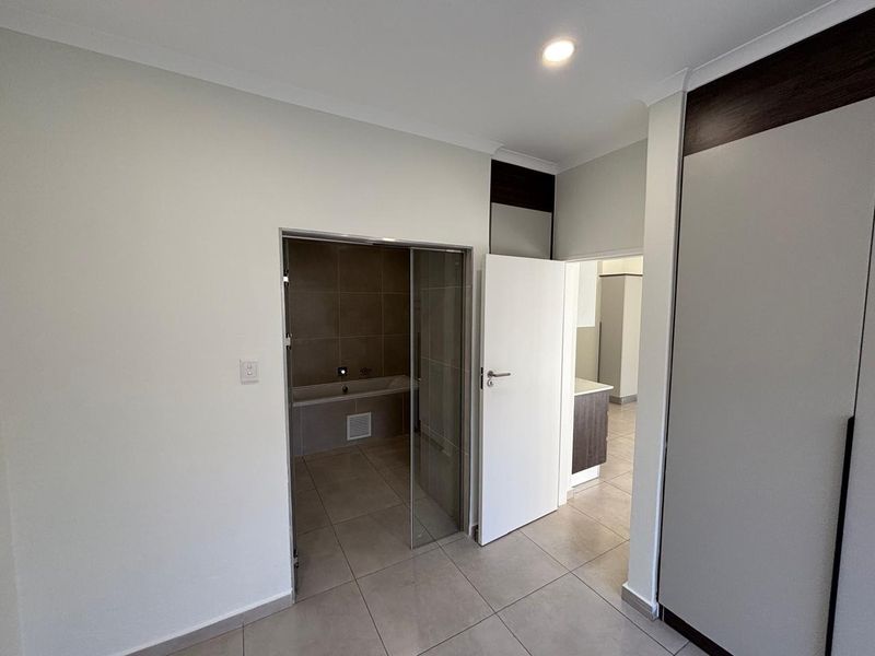 Spacious bedroom featuring neutral tiled flooring, built-in cupboards, and direct access to an en-suite bathroom with a glass door entry.