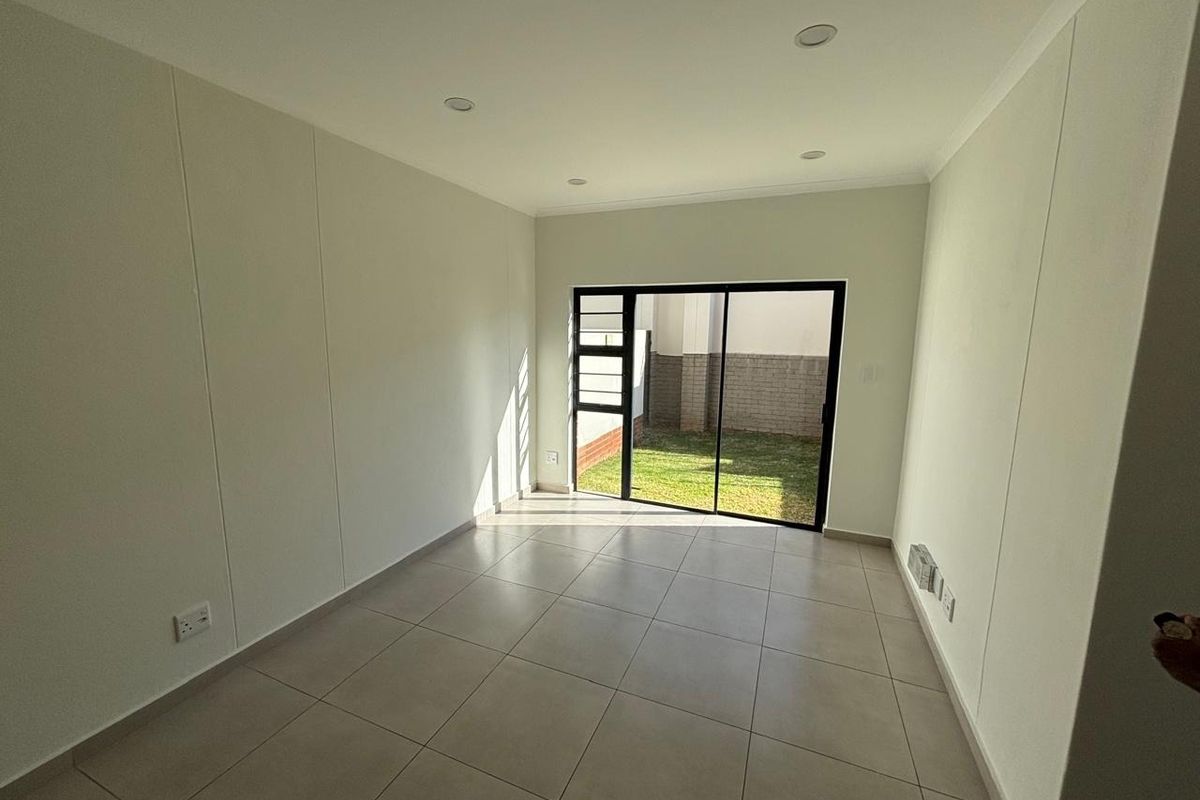 A bright, empty living room featuring neutral tiled flooring, recessed lighting, and a sliding glass door providing access to the rear garden area.