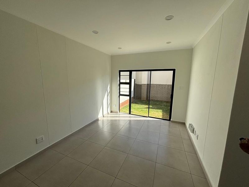 A bright, empty living room featuring neutral tiled flooring, recessed lighting, and a sliding glass door providing access to the rear garden area.