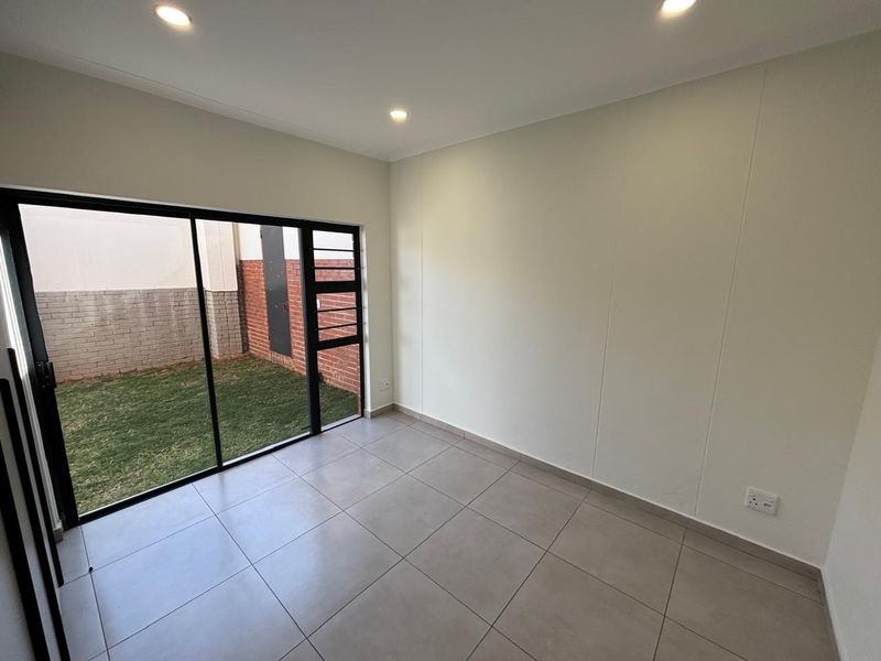 A bright, empty living room featuring neutral tiled flooring, recessed lighting, and a large sliding glass door providing access to the rear garden area.