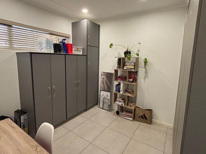 A functional office space featuring neutral tiled flooring, built-in grey cabinetry for storage, and venetian blinds for light control.