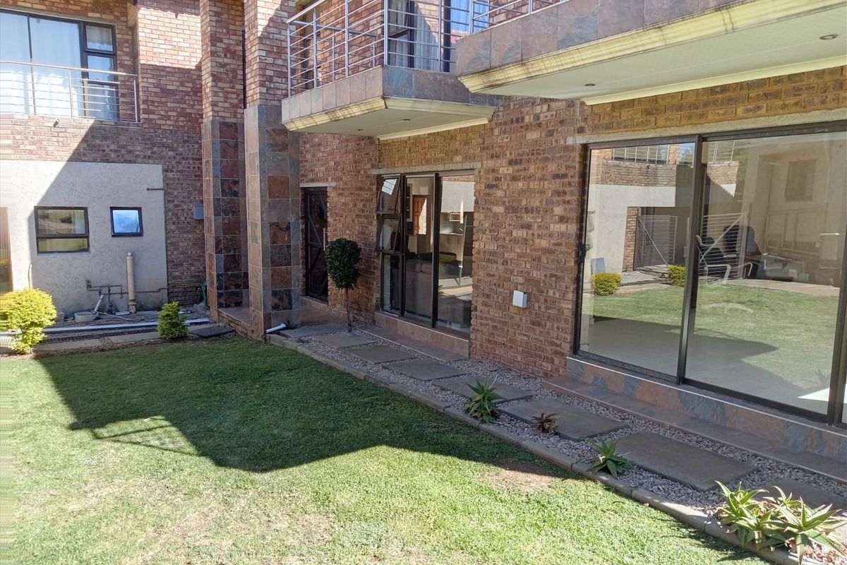 Rear view of a double-story face brick house featuring a landscaped garden, paved walkway, and an upper-level balcony with a stainless steel balustrade.