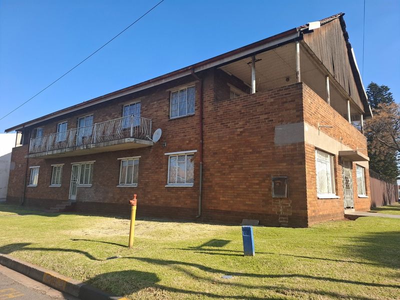 A two-story face brick residential building featuring a balcony with metal railings and security gates, set on a grassed lot.