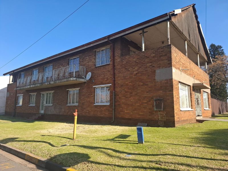 A two-story face brick residential building featuring a balcony, steel security gates, and a lawn area, showing signs of exterior wear and maintenance needs.