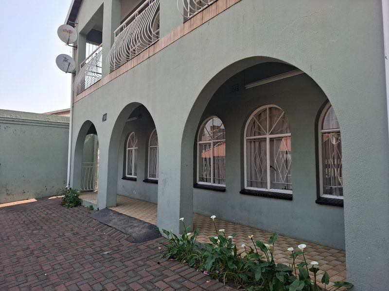 Front facade of a multi-story house featuring an arched covered walkway, paved driveway, and balcony with decorative metal railings.
