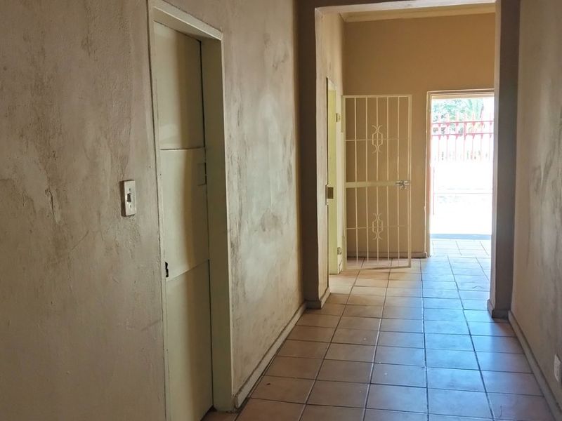 A vacant hallway featuring tiled flooring and an ornate plaster ceiling, leading towards an exterior doorway with a security gate.