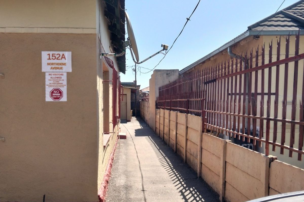 A narrow exterior side passage between two residential buildings featuring concrete boundary walls, security fencing, and a paved walkway.