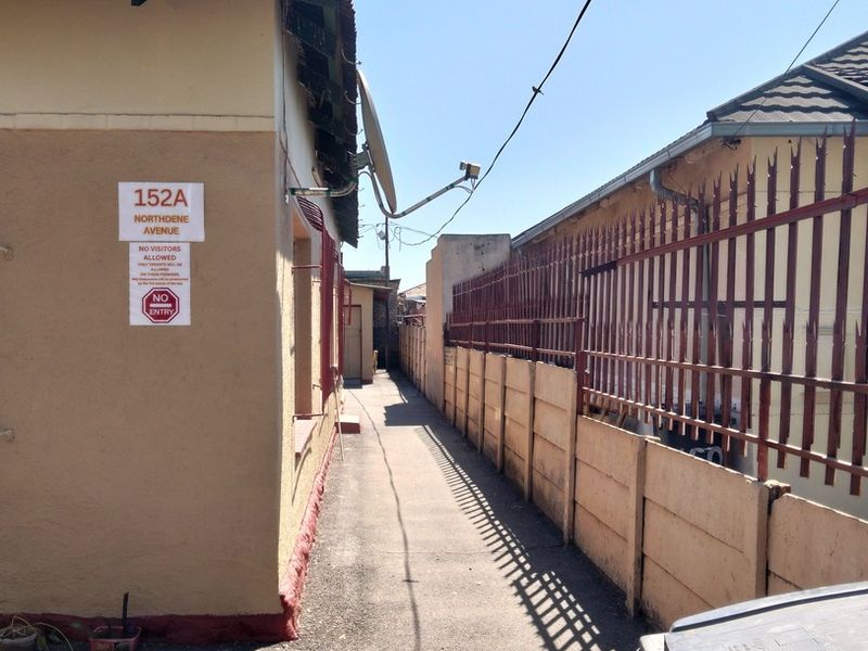 A narrow exterior side passage between two residential buildings featuring concrete boundary walls, security fencing, and a paved walkway.
