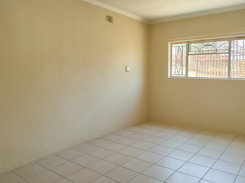 A vacant bedroom featuring neutral tiled flooring, plain walls, and a window fitted with security bars.