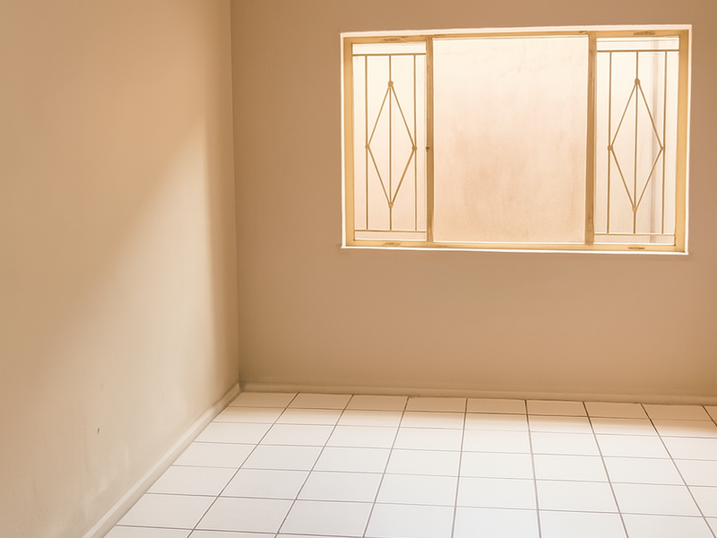 A clean, empty bedroom featuring neutral wall paint, white tiled flooring, and a window fitted with security burglar bars.