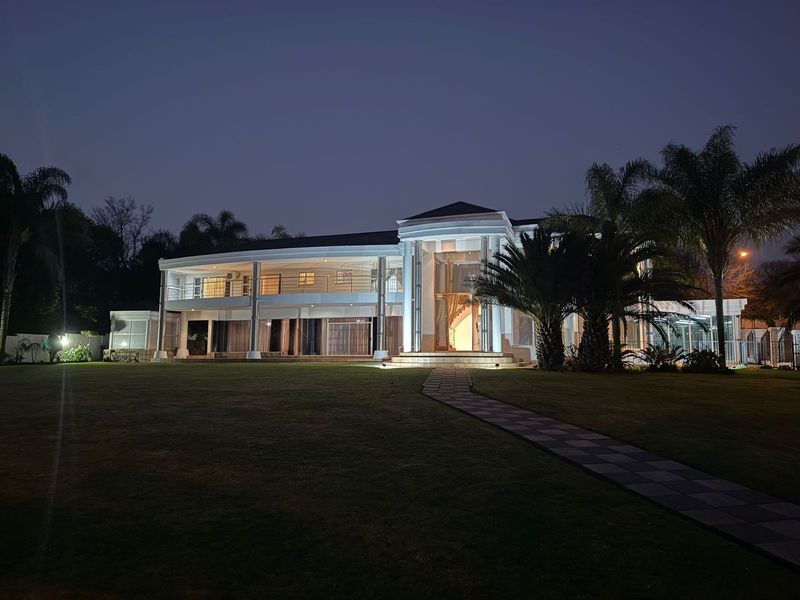 A grand two-story residence at dusk, featuring a prominent columned entrance, expansive covered balcony, and a well-manicured lawn with a paved walkway.