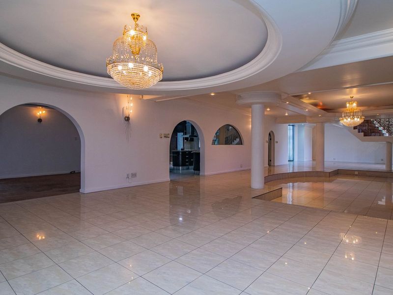 Spacious open-plan living area featuring tiled floors, decorative recessed ceilings with chandeliers, arched architectural details, and a sunken floor section with a staircase.