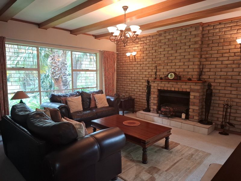 Spacious living room featuring a prominent brick fireplace, exposed timber ceiling beams, and large windows providing natural light and garden views.