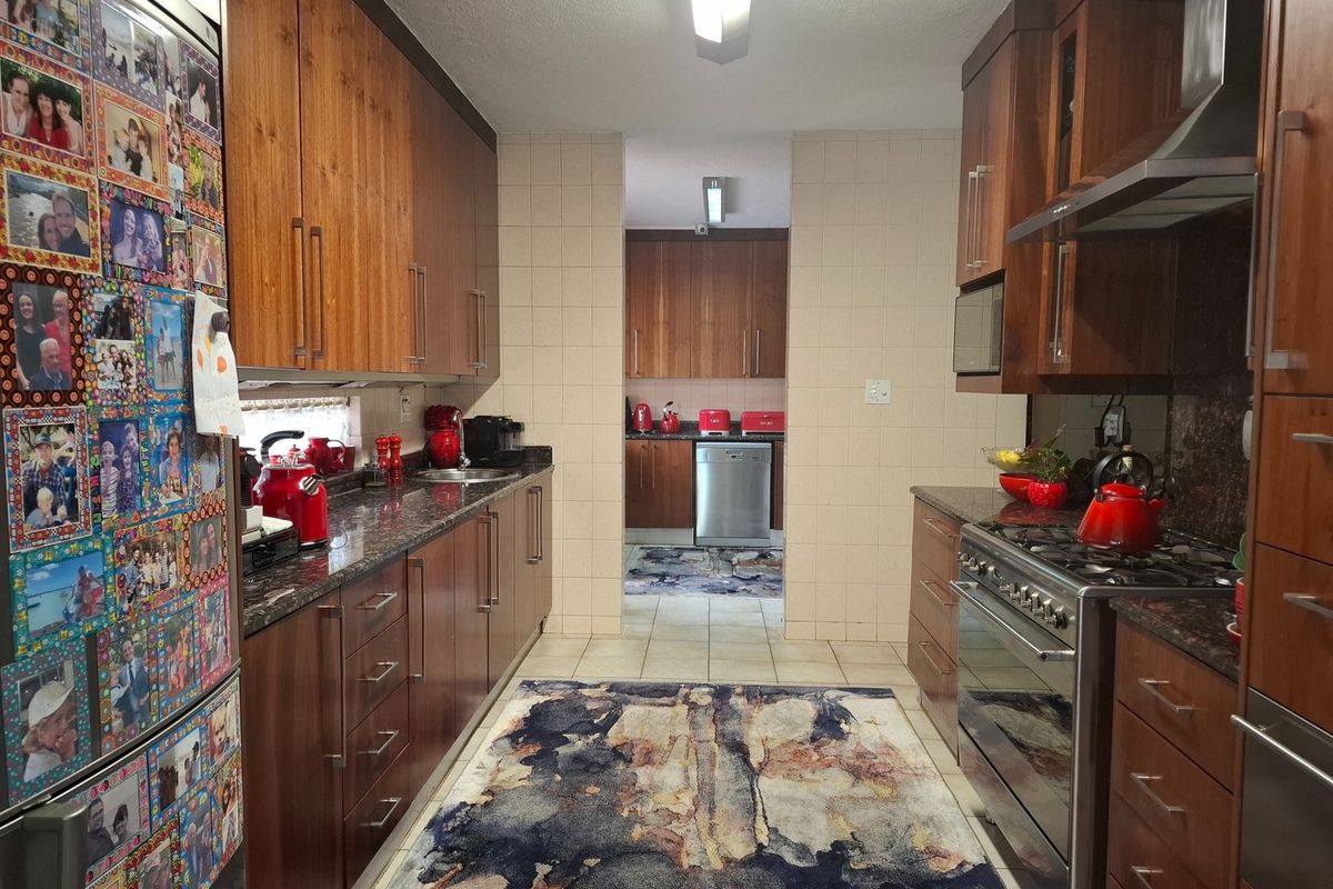 A functional galley-style kitchen featuring warm wooden cabinetry, granite countertops, stainless steel appliances, and a tiled backsplash, leading into a secondary scullery area.