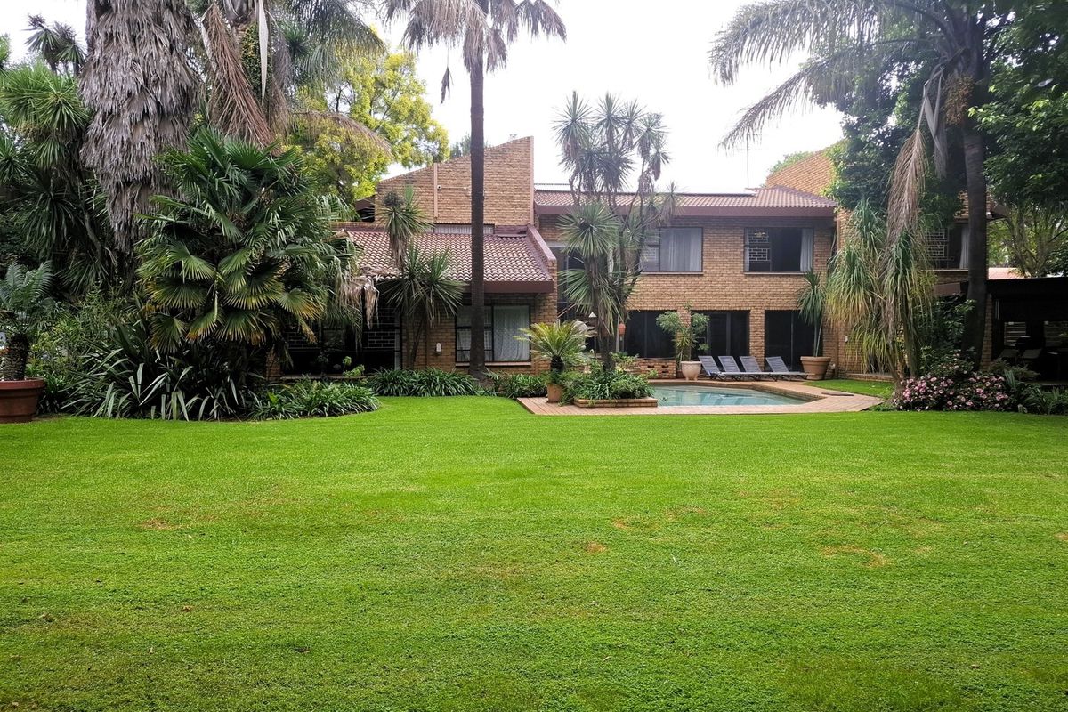 A spacious rear garden featuring a well-maintained lawn, mature trees, and a swimming pool set against a classic brick residential facade.