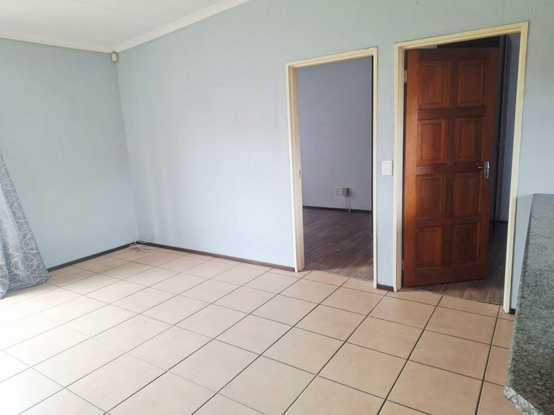An empty living area featuring neutral tiled flooring, a solid wooden door, and access to an adjacent room.