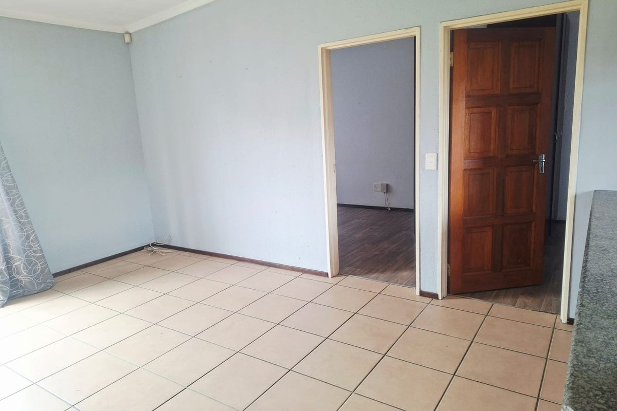An empty living area featuring neutral tiled flooring, a solid wooden door, and access to an adjacent room.
