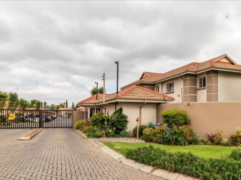 The entrance to the apartment complex features a secure gated access point, paved driveway, and well-maintained perimeter walls with electric fencing.
