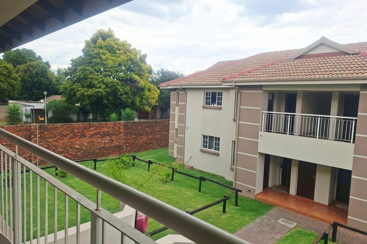 View of the apartment complex exterior featuring a tiled roof, balcony, paved walkway, and a well-maintained communal garden area with brick boundary walls.