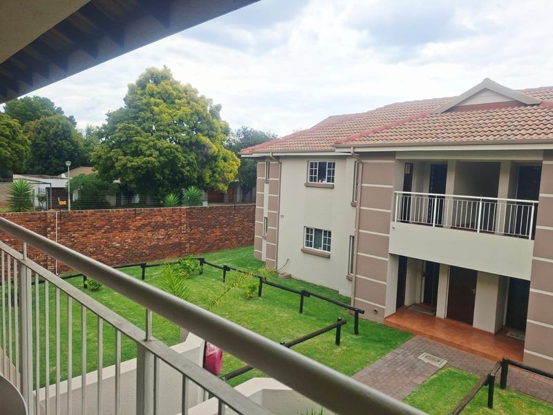 View of the apartment complex exterior featuring a tiled roof, balcony, paved walkway, and a well-maintained communal garden area with brick boundary walls.