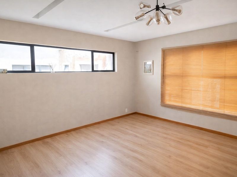 A bright and spacious bedroom featuring light laminate flooring, modern lighting, and large windows with timber venetian blinds.