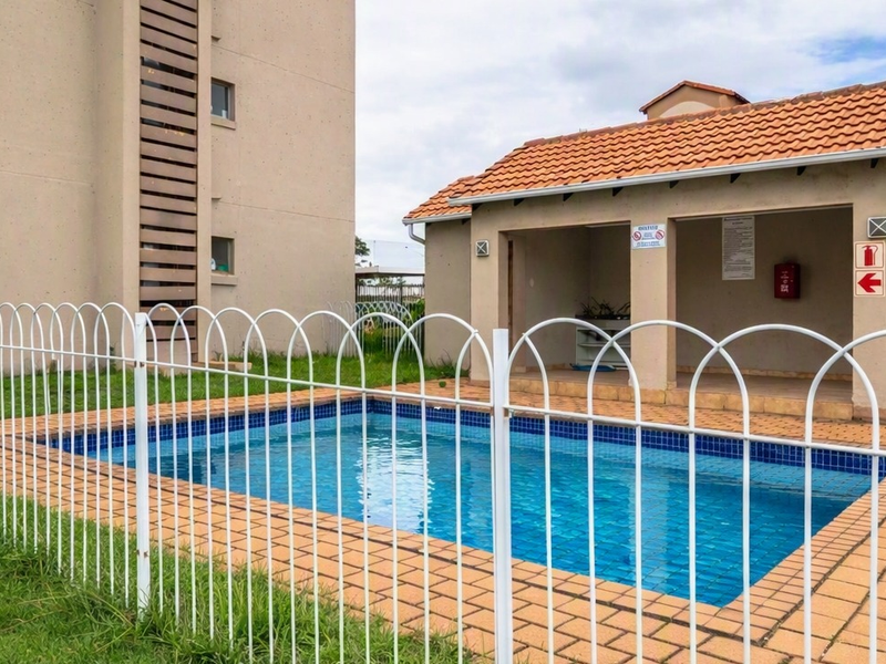 Communal swimming pool area featuring a paved surround, safety fencing, and an adjacent covered patio structure with tiled roofing.