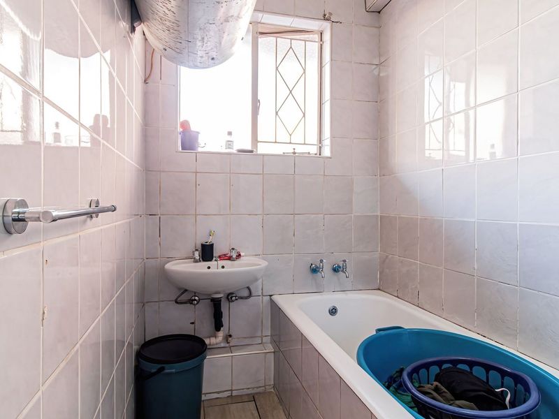 A compact bathroom featuring a built-in bathtub, wall-mounted basin, and full-height ceramic wall tiling, requiring cosmetic updates to improve its overall condition.