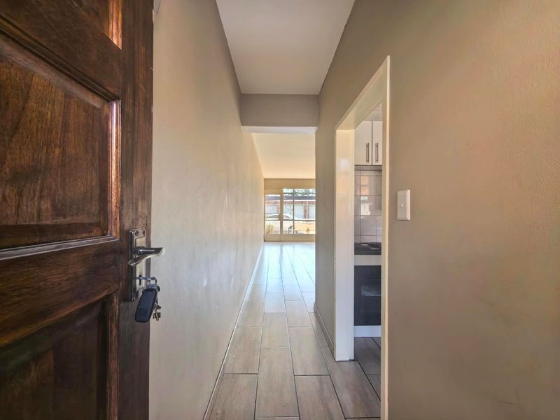 A clean, neutral-toned hallway featuring tiled flooring and a solid wood entrance door, providing access to the kitchen and main living area.
