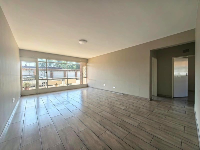 A spacious, empty living room featuring wood-look tiled flooring and a large window wall providing ample natural light.