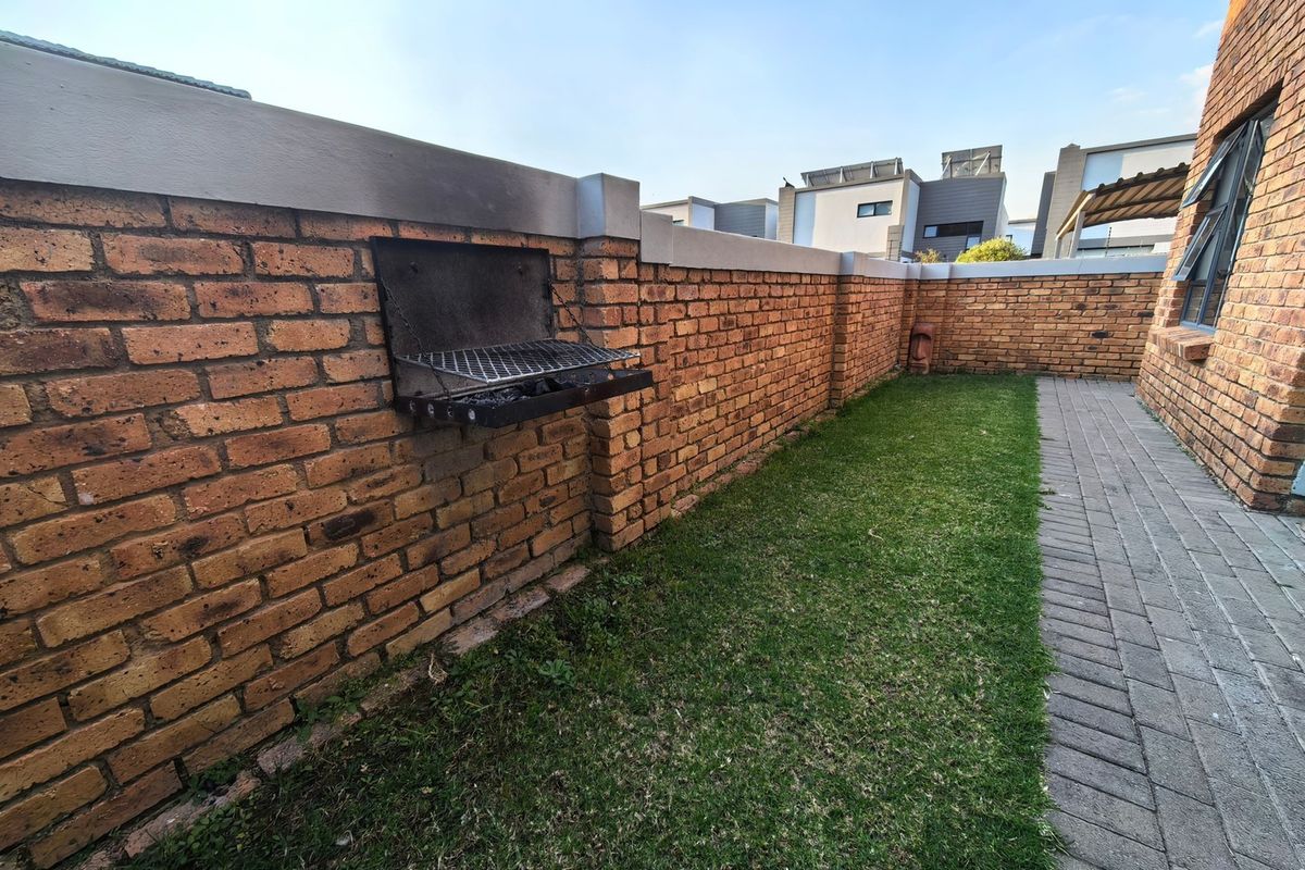 A private rear garden area featuring a brick boundary wall with a built-in braai, a paved walkway, and a small lawn.
