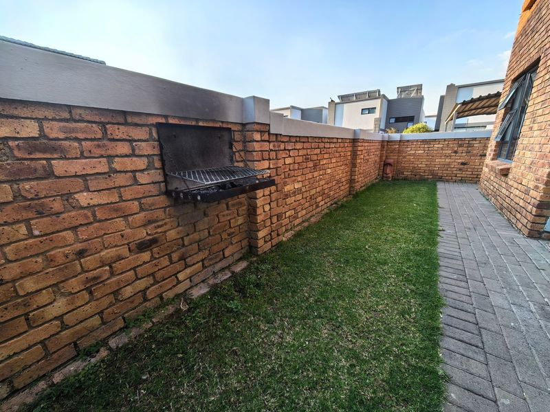 A private rear garden area featuring a brick boundary wall with a built-in braai, a paved walkway, and a small lawn.