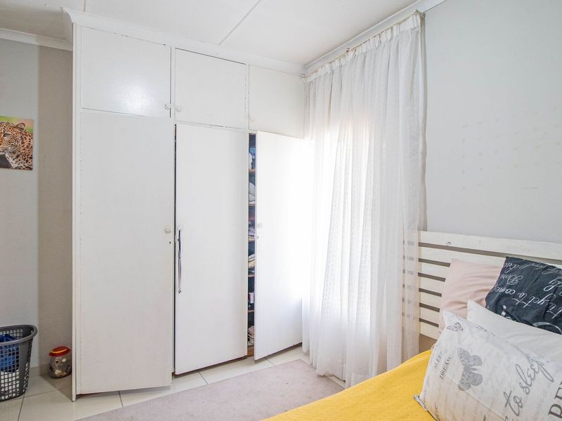 A bedroom featuring white built-in cupboards, tiled flooring, and a window with sheer curtains.
