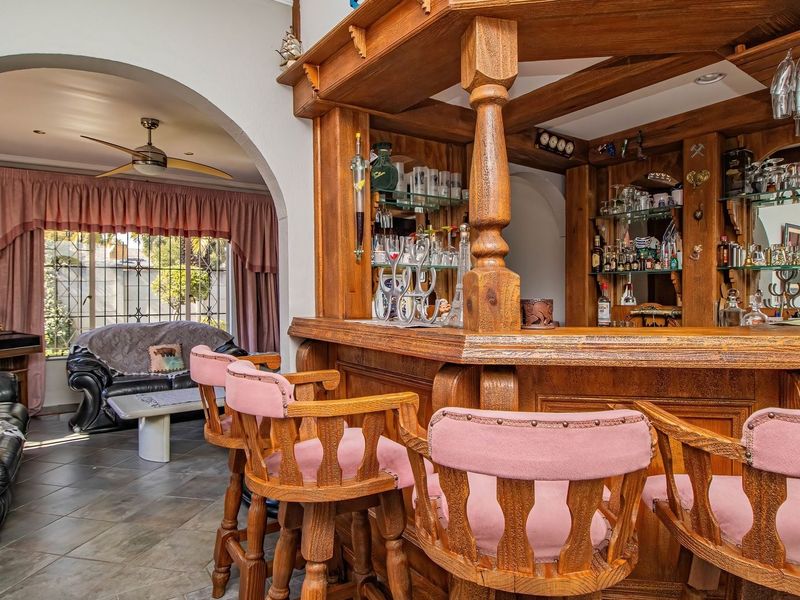 A dedicated home bar area featuring a substantial built-in wooden counter, glass shelving, and tiled flooring, connected to an adjacent living space via an archway.