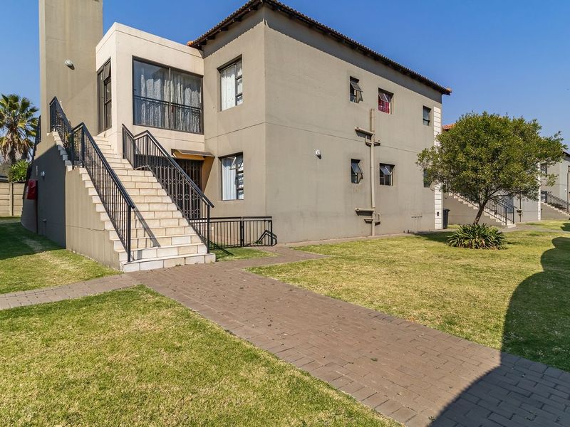 Exterior view of an apartment building featuring a paved walkway, external staircase with metal railings, and adjacent carport parking.