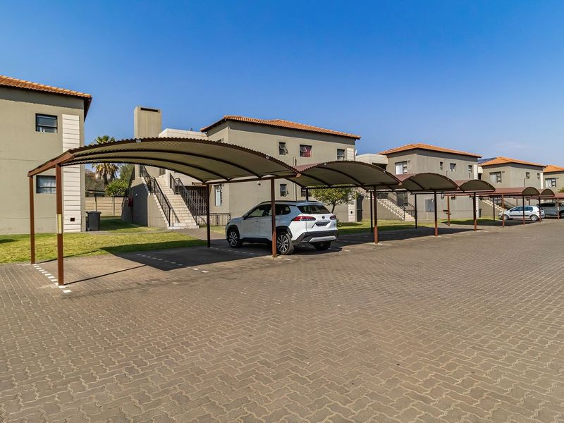 Exterior view of an apartment complex featuring covered parking bays, paved driveway, and multi-story residential buildings with tiled roofs.